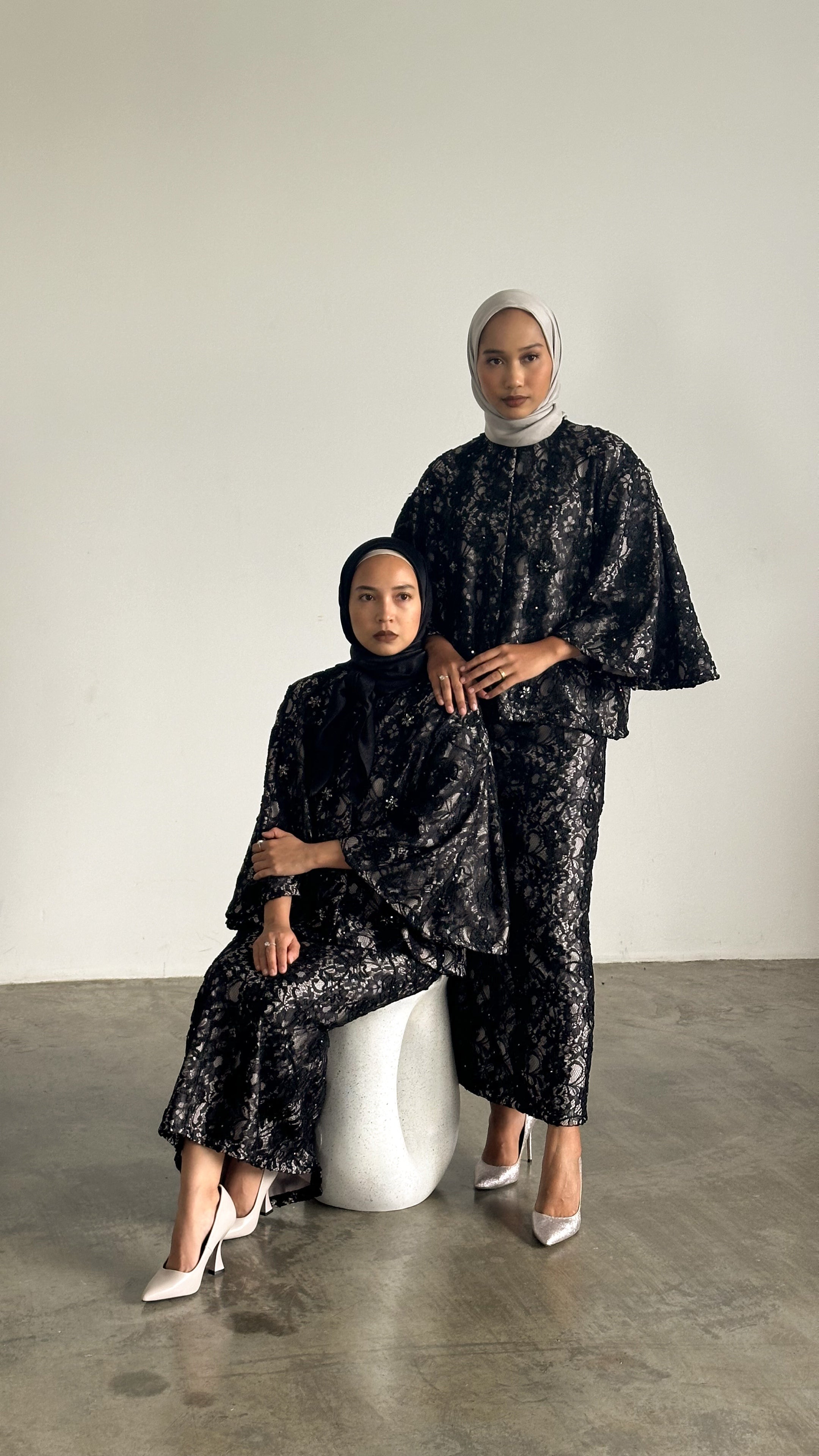 Two women in matching black patterned outfits sitting on a white stool against a plain background.